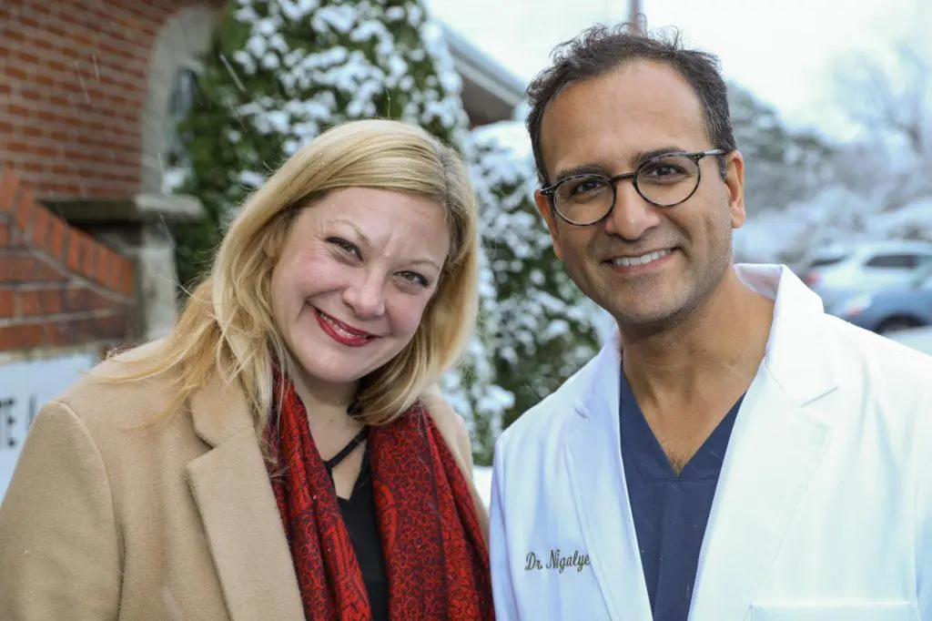 A woman and a man, both smiling, standing outdoors in front of snowy trees. The woman has blonde hair, wearing a tan coat and red scarf. The man has glasses, short dark hair, wearing a white lab coat with name tag, and dark scrubs underneath.