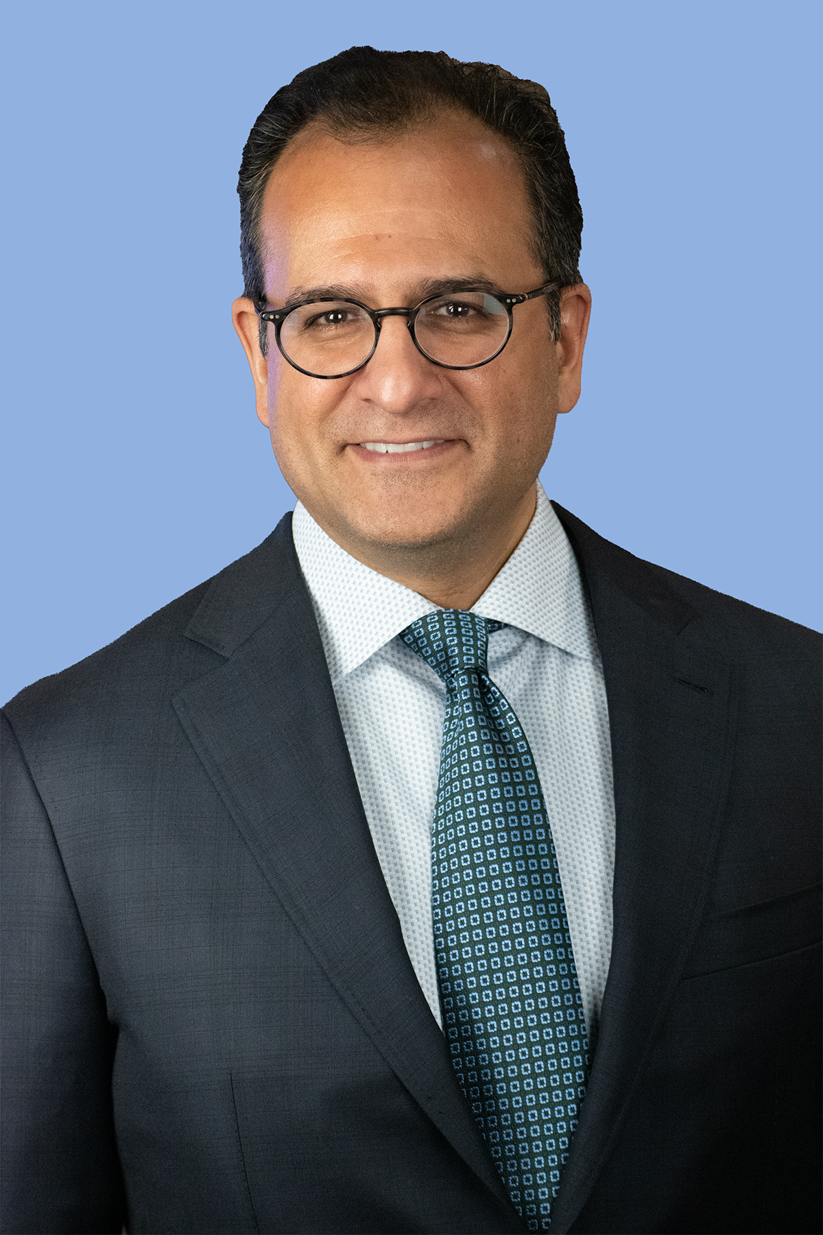 Professional headshot of Dr. Sanil B. Nigalye, DDS, MD, of Niagara Dental Implant & Oral Surgery, with glasses, wearing a dark suit, white shirt, and a patterned blue tie, against a light blue background.
