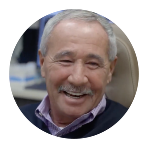 A smiling elderly man with gray hair and a mustache, wearing a collared shirt and sweater, sitting in a chair.