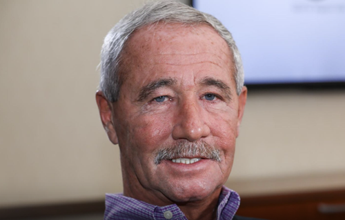 Close-up photo of an older man with gray hair and a mustache, smiling, wearing a purple collared shirt, in an indoor setting.