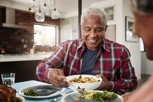 An elderly man in a red plaid shirt smiling as he serves himself a bowl of food at a table in a modern kitchen.