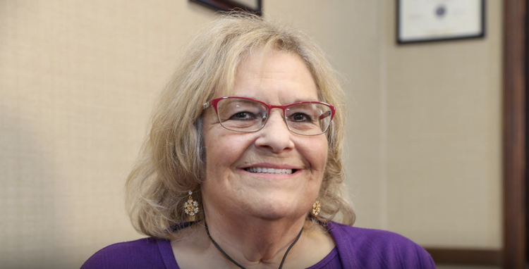 A smiling woman with blonde hair, wearing red glasses, earrings, and a purple top in an office setting.
