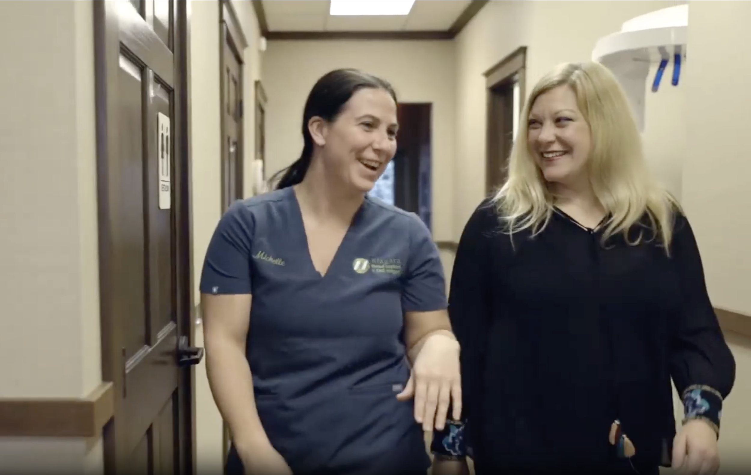 Two women walking down a hallway, smiling and conversing, one wearing scrubs and the other in casual black clothing.