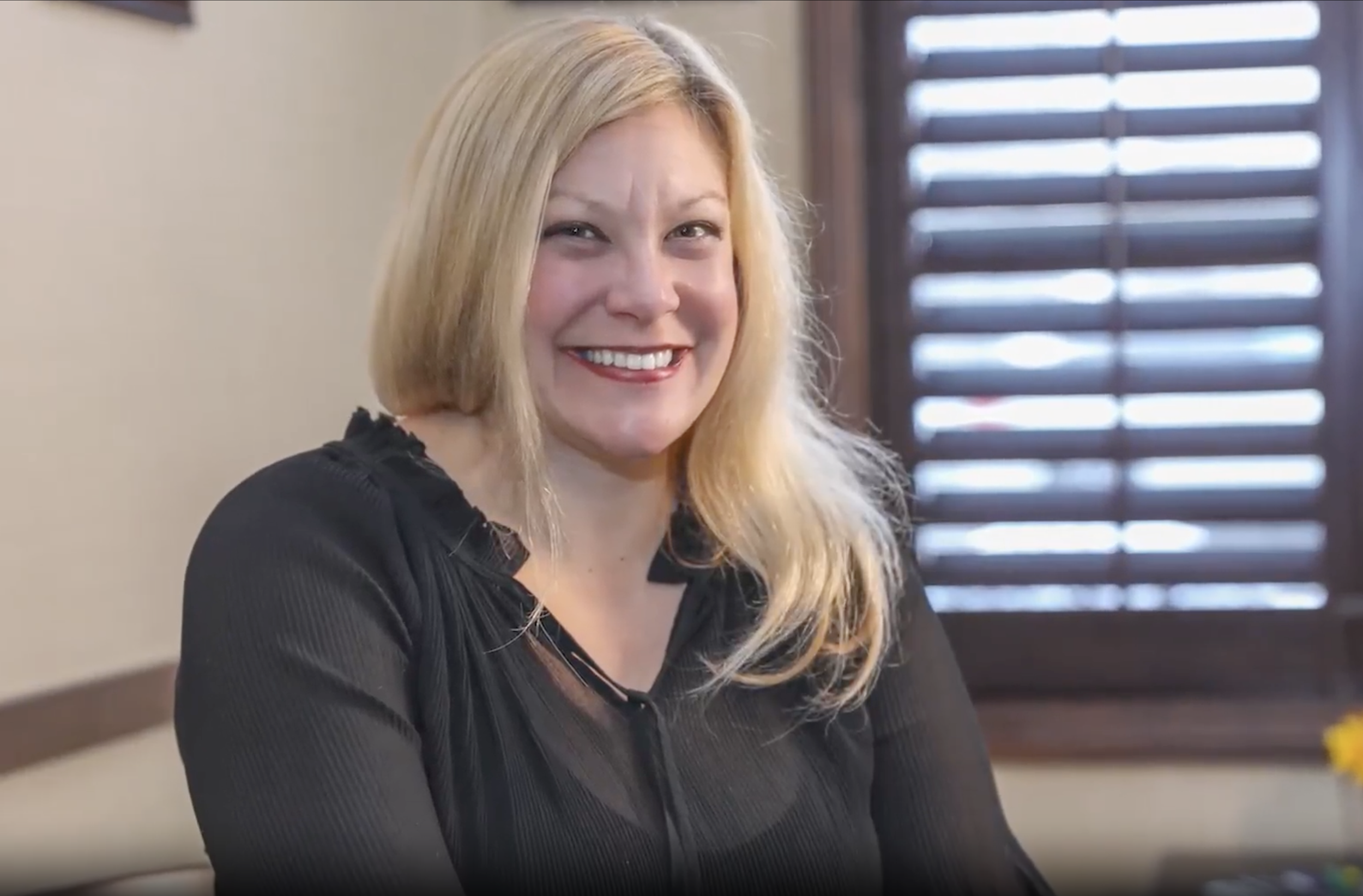 A woman with long blonde hair, smiling and wearing a black top, sitting indoors near a window with wooden shutters.