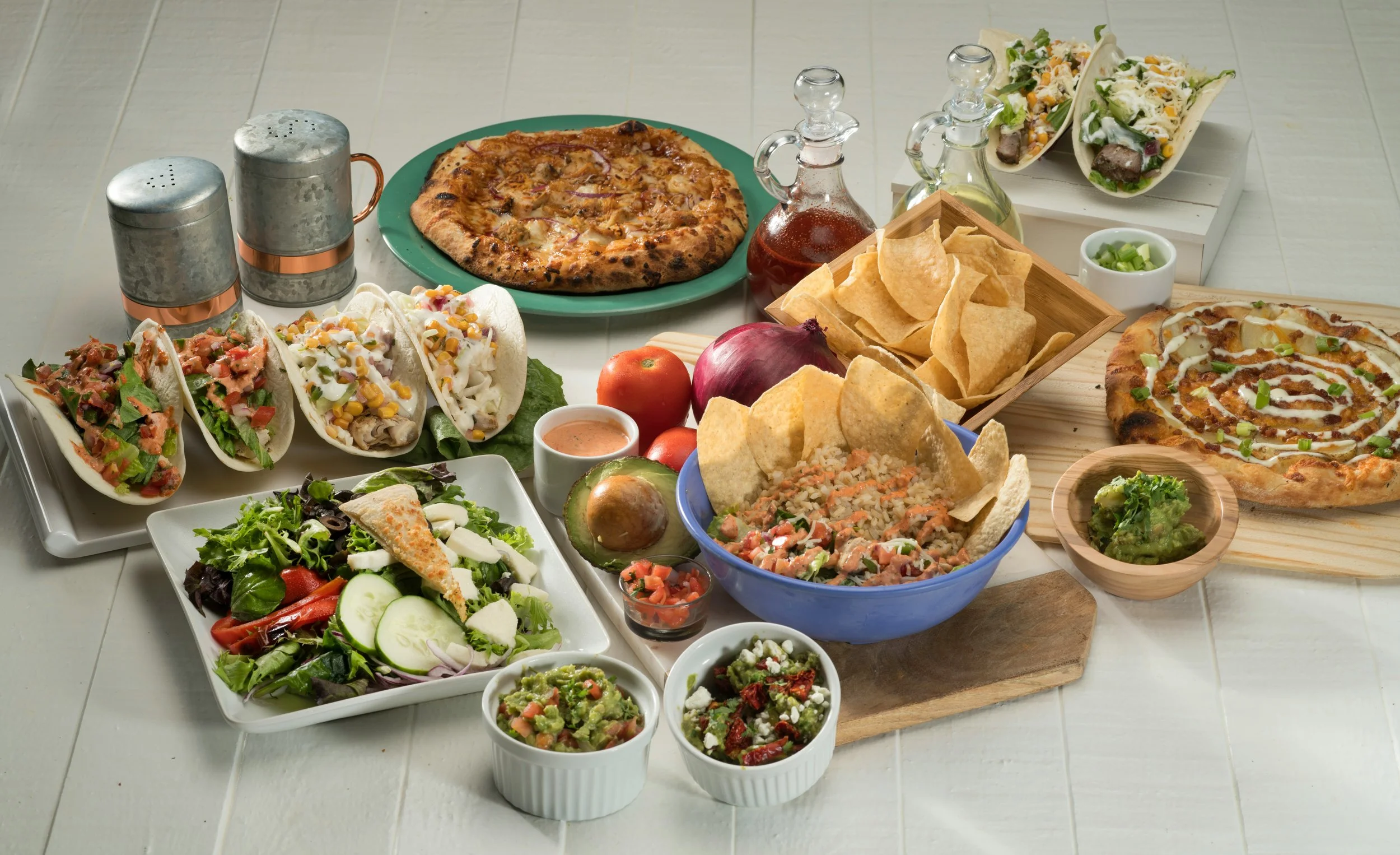 Assorted Mexican dishes including tacos, two pizzas, nachos, tacos, salad, rice, guacamole, salsa, tomato, onion, sauces, and beverages on a table.